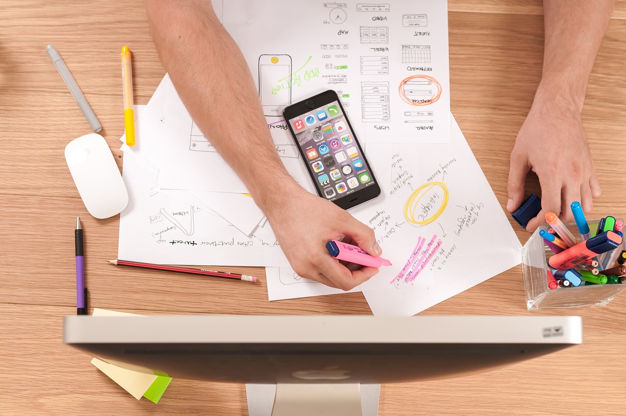 Person working on a project, surrounded by colored markers, sticky notes, and papers with design sketches and mobile app ideas.