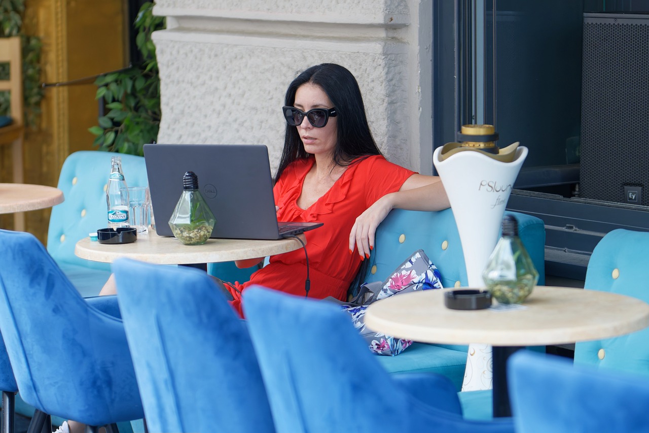 Woman in sunglasses relaxing at a café, using a laptop on a sunny day.