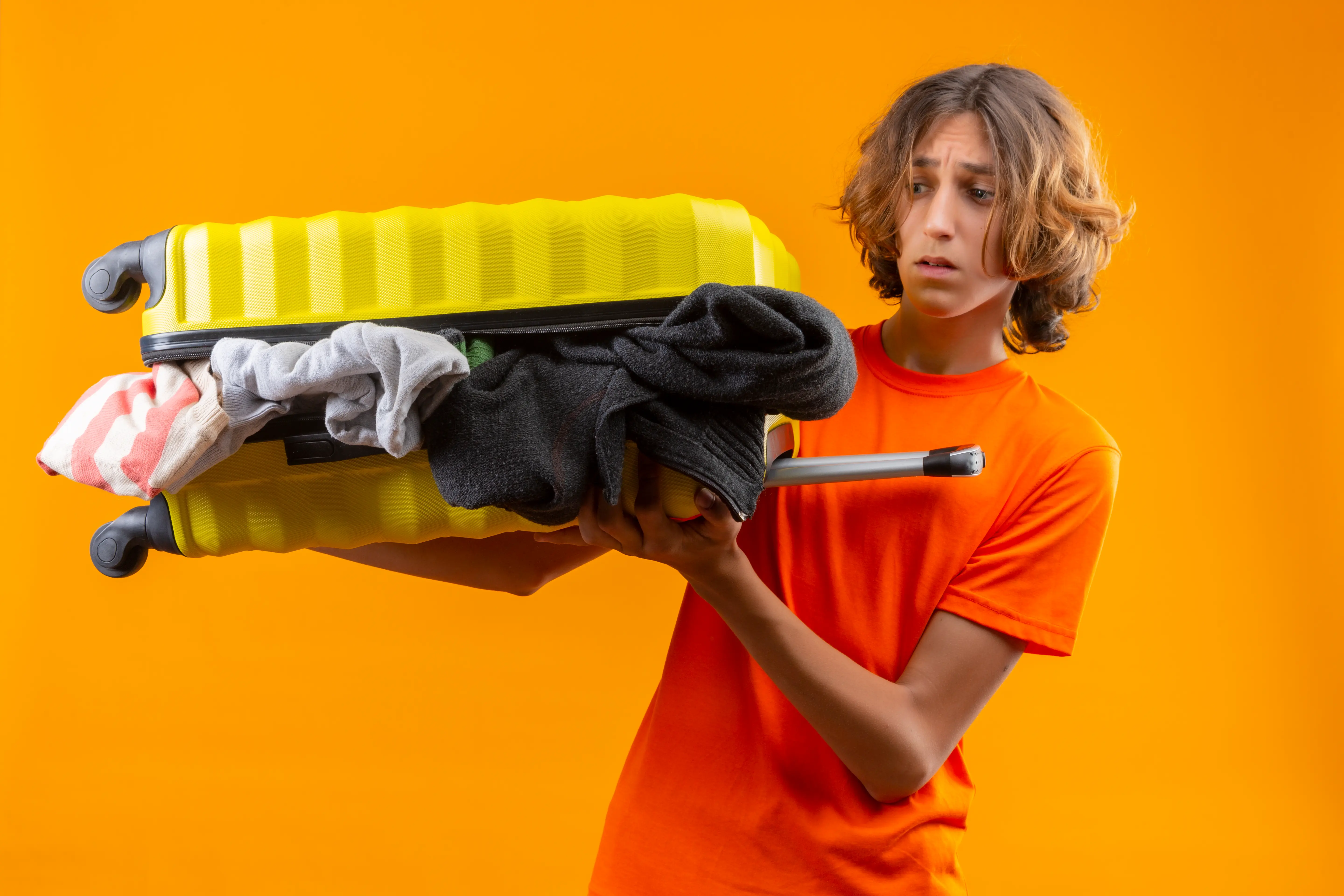 Young traveler holding an overstuffed yellow suitcase with clothes spilling out, symbolizing packing mistakes